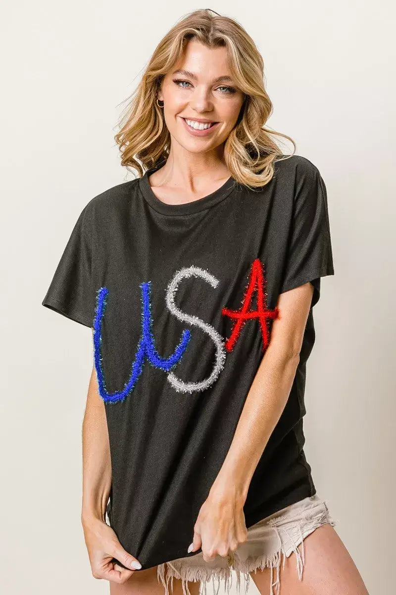 Woman wearing a black t-shirt featuring colorful rhinestone 'USA' letters, styled with casual shorts.