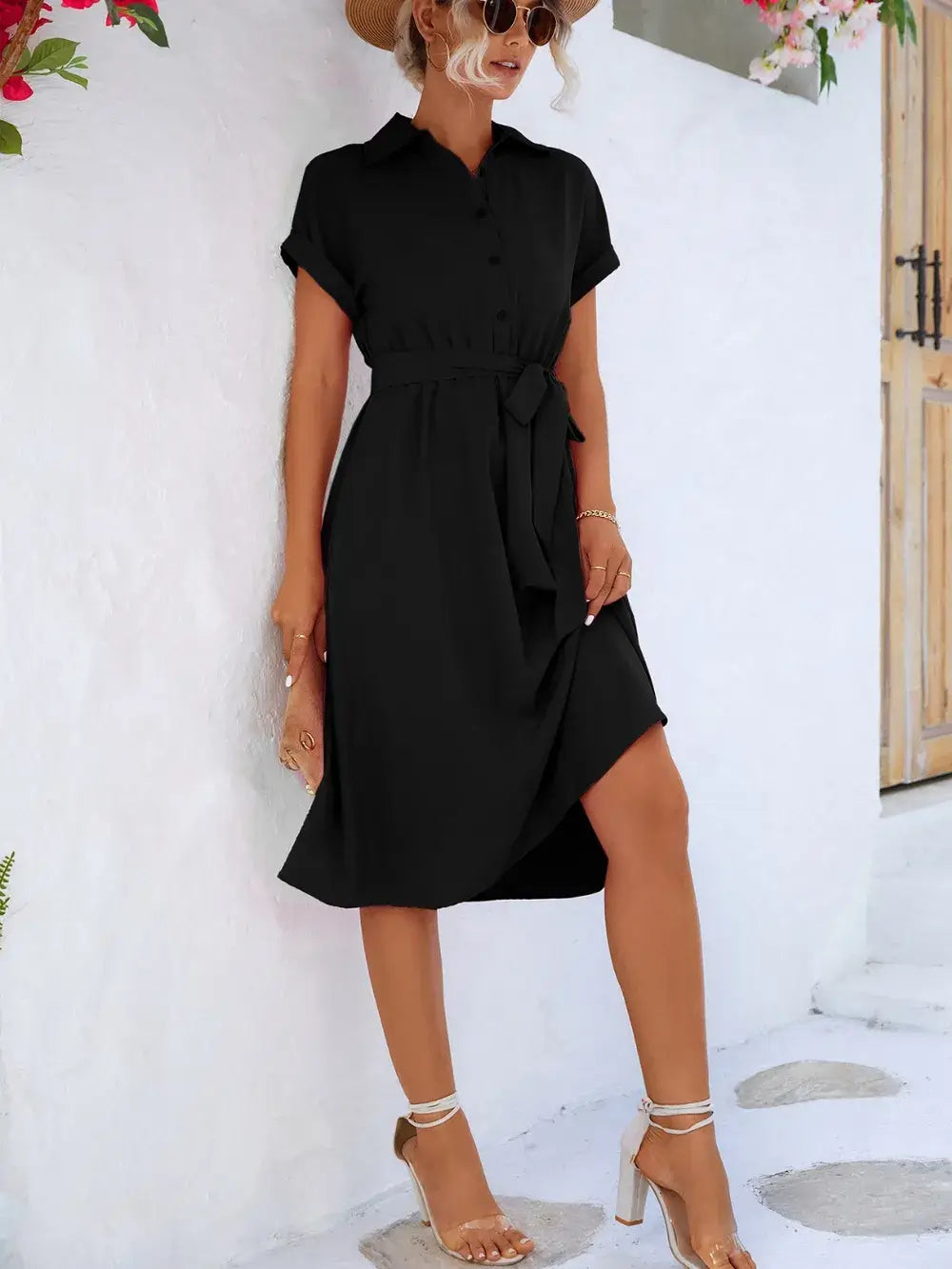 Model wearing a black Buttoned Tie Waist Short Sleeve Dress with a bow, standing against a white wall.