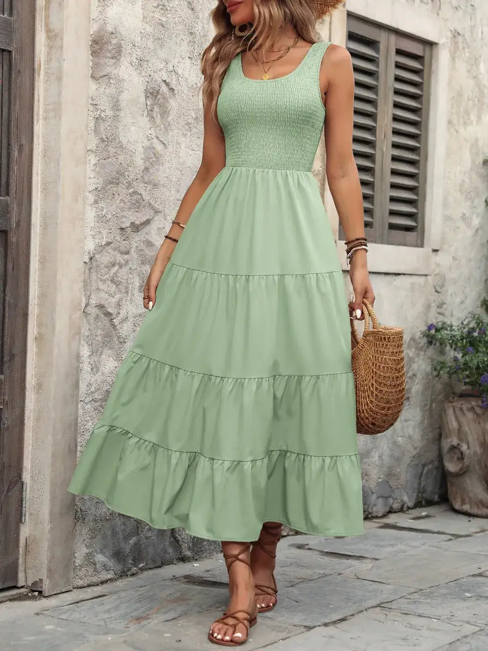 Woman wearing Ivy Lane Smocked Scoop Neck Sleeveless Tank Dress in green, standing on a stone pathway, holding a straw bag.