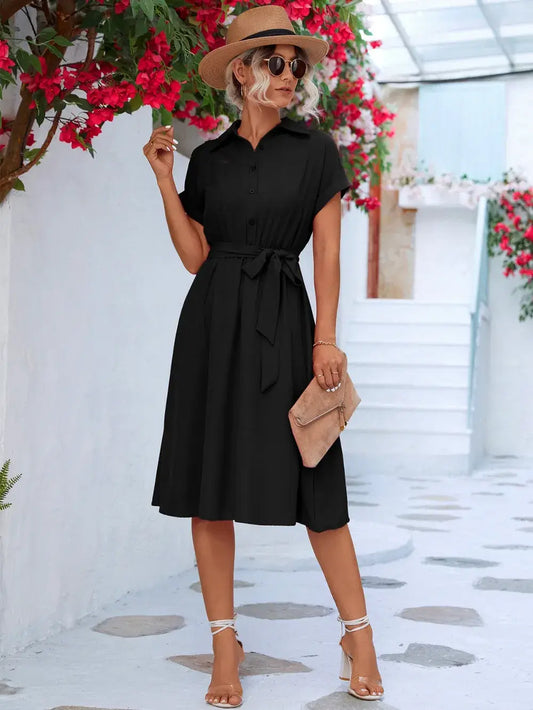 Model wearing a black buttoned tie waist short sleeve dress, styled with a hat, sunglasses, and a clutch.