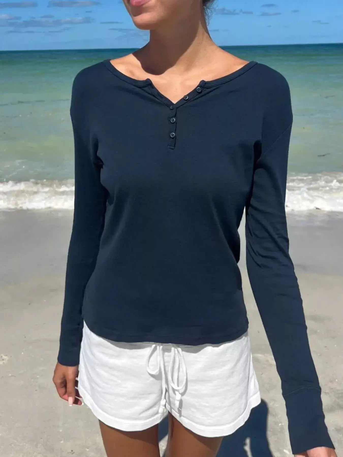 Model wearing a navy striped long sleeve henley T-shirt and white shorts on a beach.