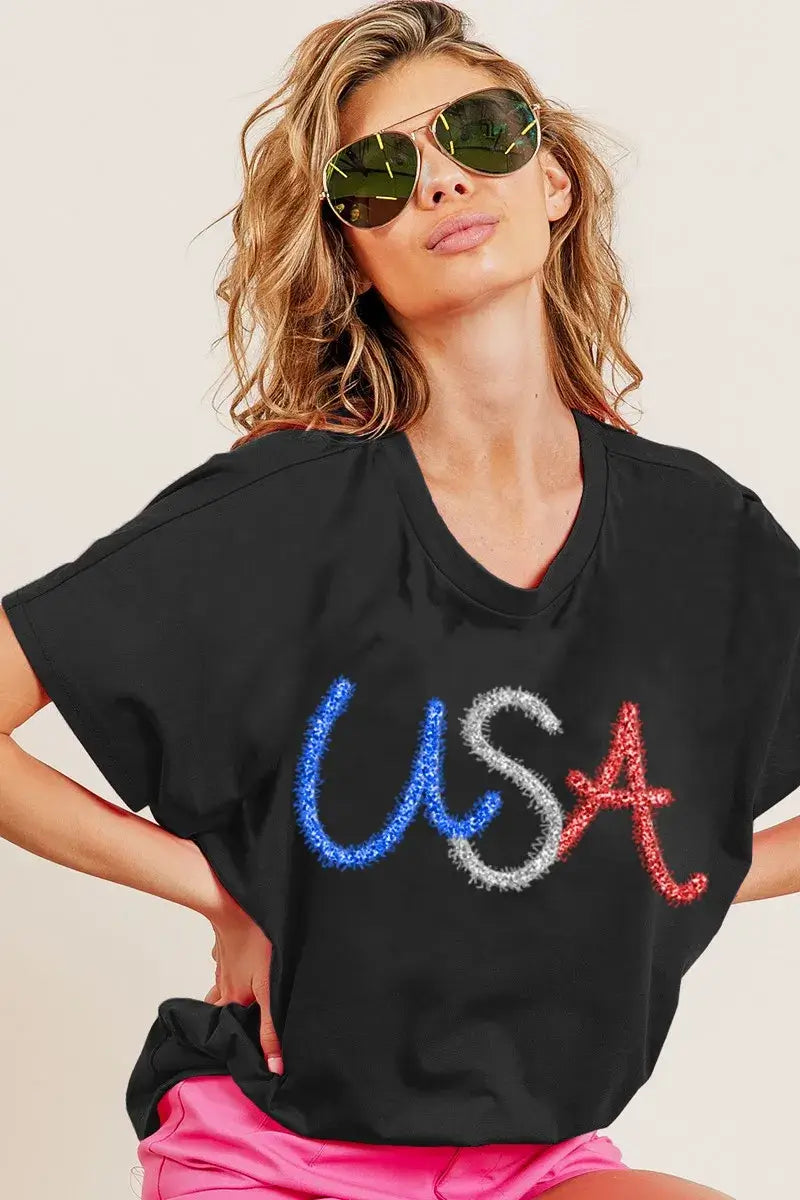 Model wearing a black oversized top with 'USA' in rhinestones and sunglasses, styled for a chic summer look.