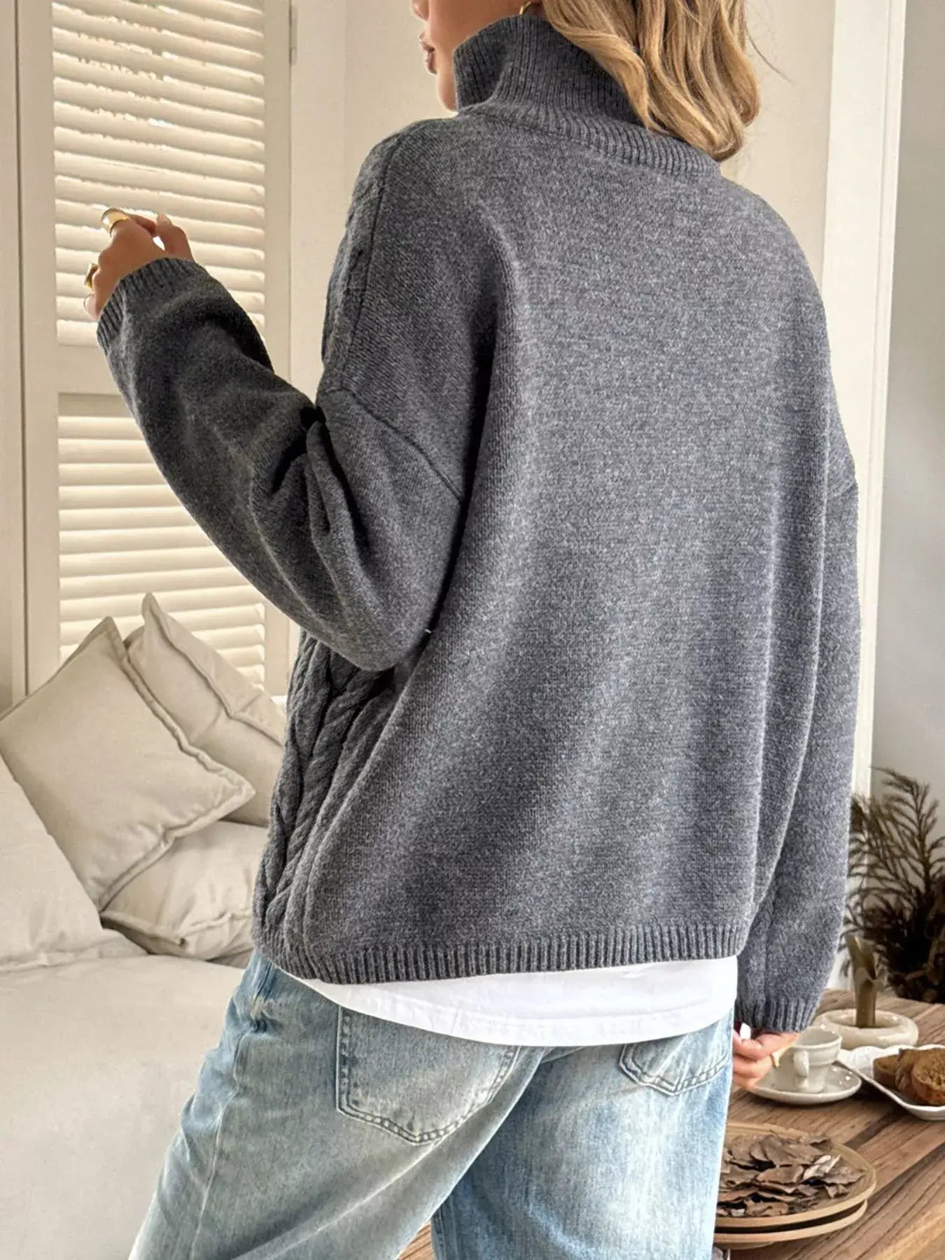 Woman wearing a zip collar cable-knit sweater in grey, showcasing the back and zip detail, paired with denim jeans.