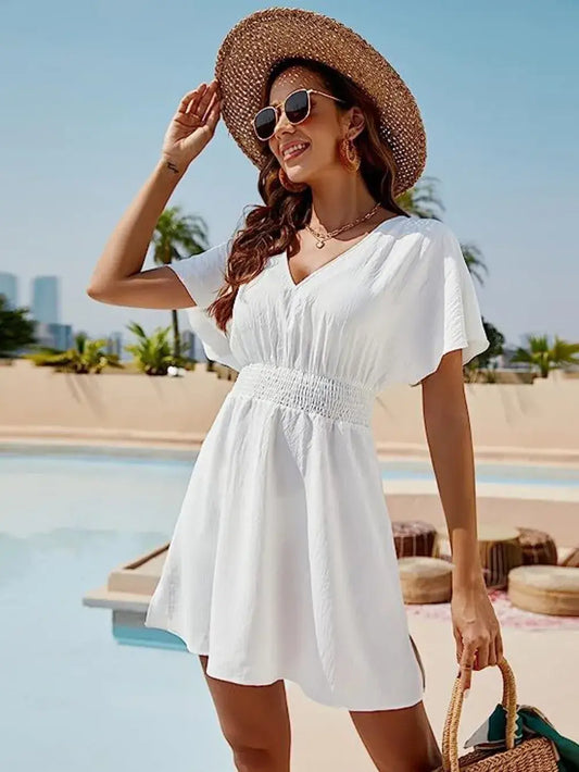 Woman in a white smocked V-neck short sleeve dress and hat, posing by a pool in a sunny setting.