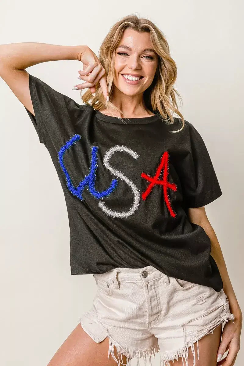 Model wearing a BiBi Metallic Letter Short Dolman Sleeve Top with 'USA' in colorful rhinestones, styled with shorts.