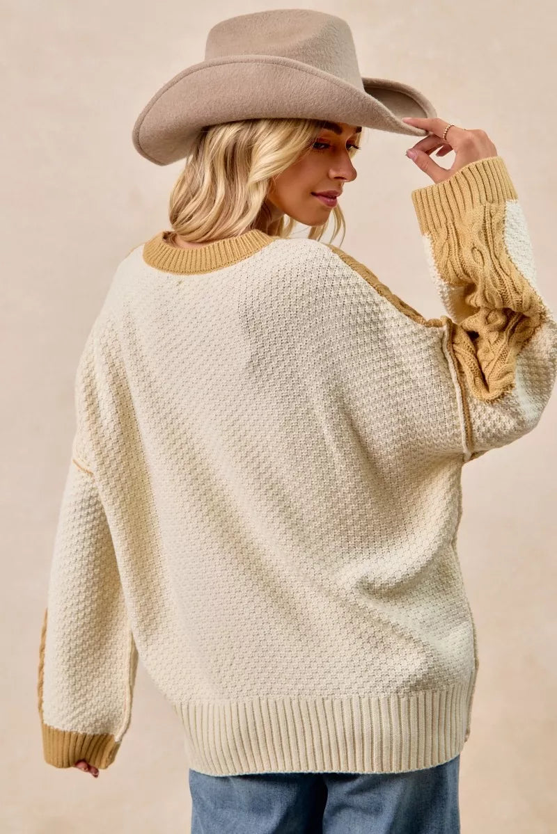 Woman wearing BiBi Cable Knit Hacci Combo Sweater with uneven hem and wide-brimmed hat, showcasing cozy style.