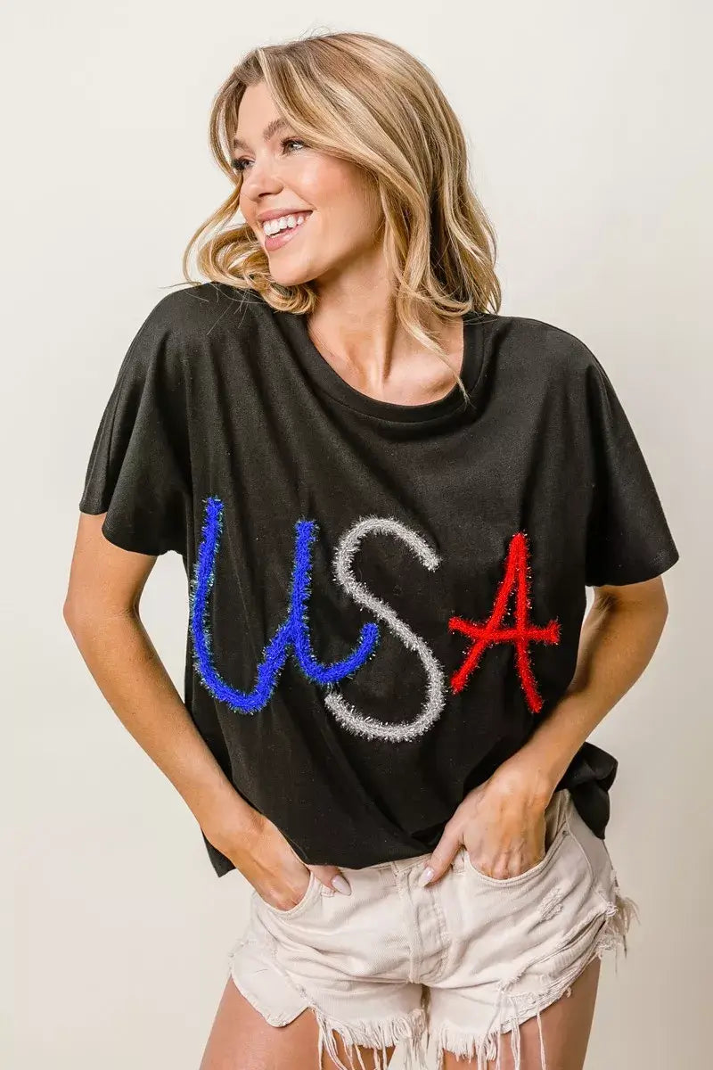 Woman wearing a black dolman sleeve top with USA letters in metallic red, white, and blue, styled with shorts.