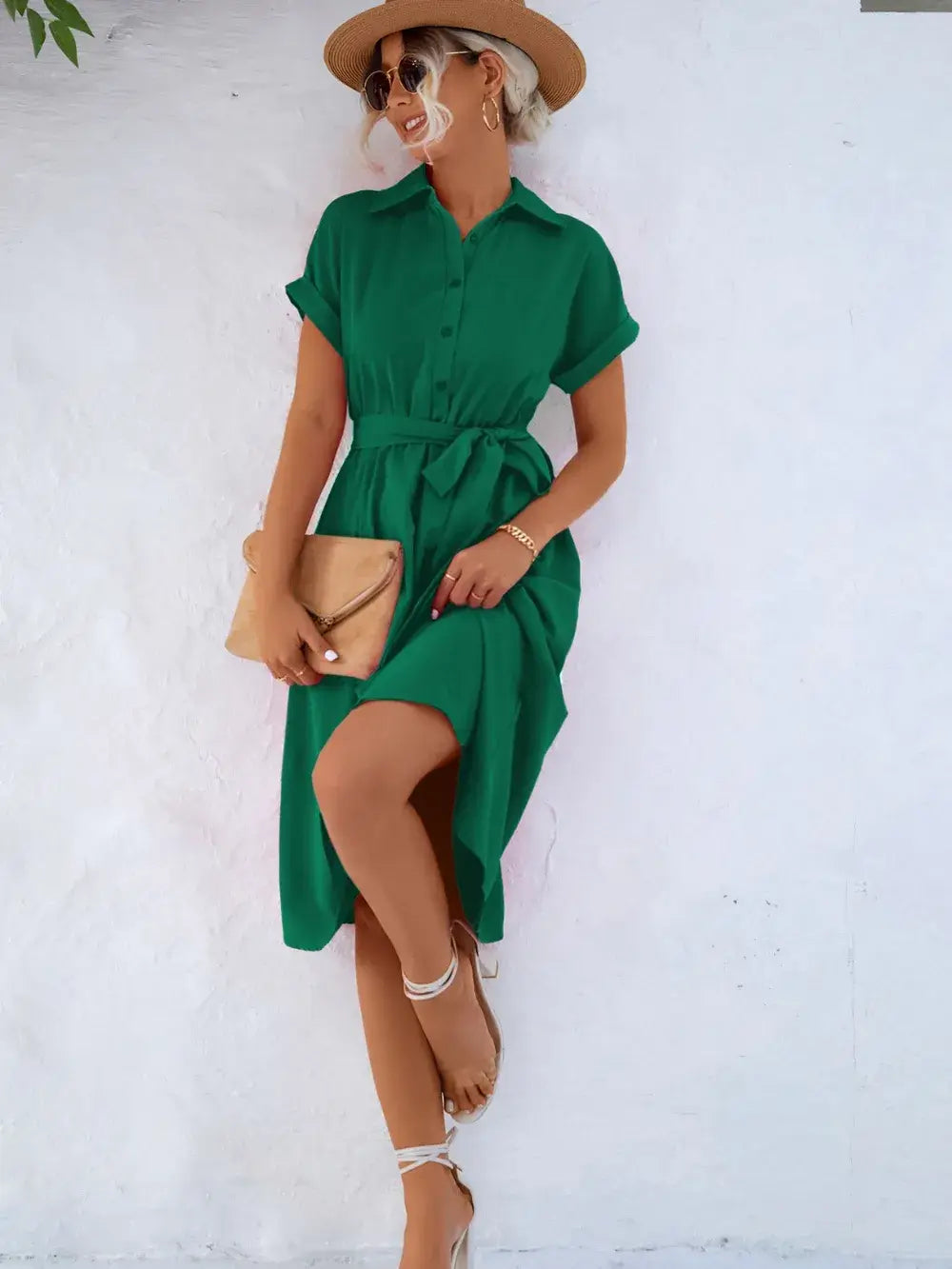 Model in a green buttoned tie waist short sleeve dress posing against a white wall, styled with a hat and clutch.