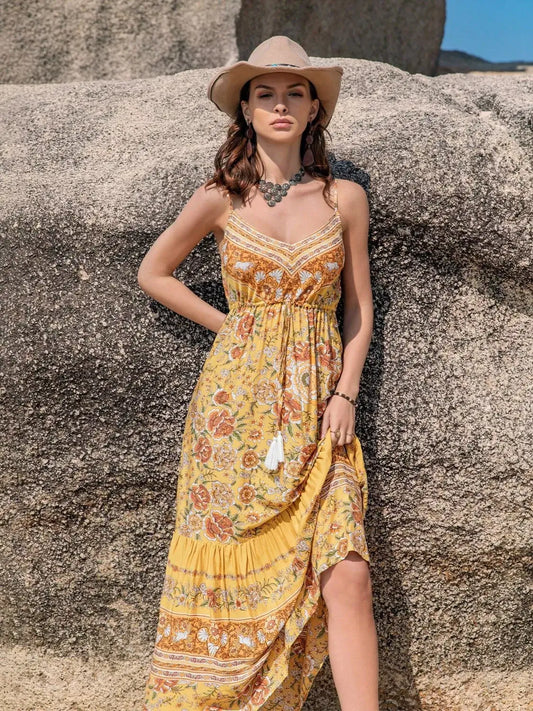 Model in a yellow floral dress posing against rocks in a summer setting, showcasing the Tassel Printed Tiered Cami Dress.