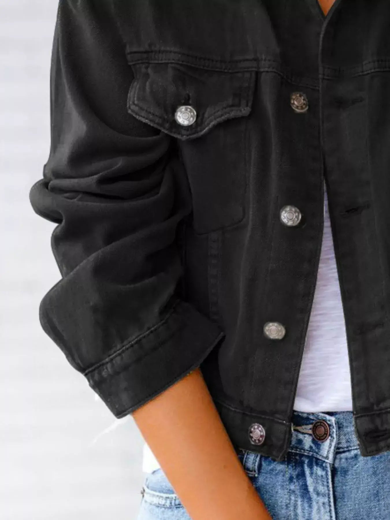 Close-up of a women's button-up black denim jacket with rolled sleeves and silver buttons, styled with jeans.