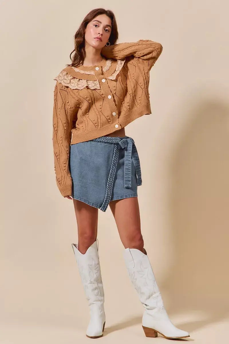 Woman in trendy wrap denim skort and knitted sweater with lace collar, styled with white cowboy boots.