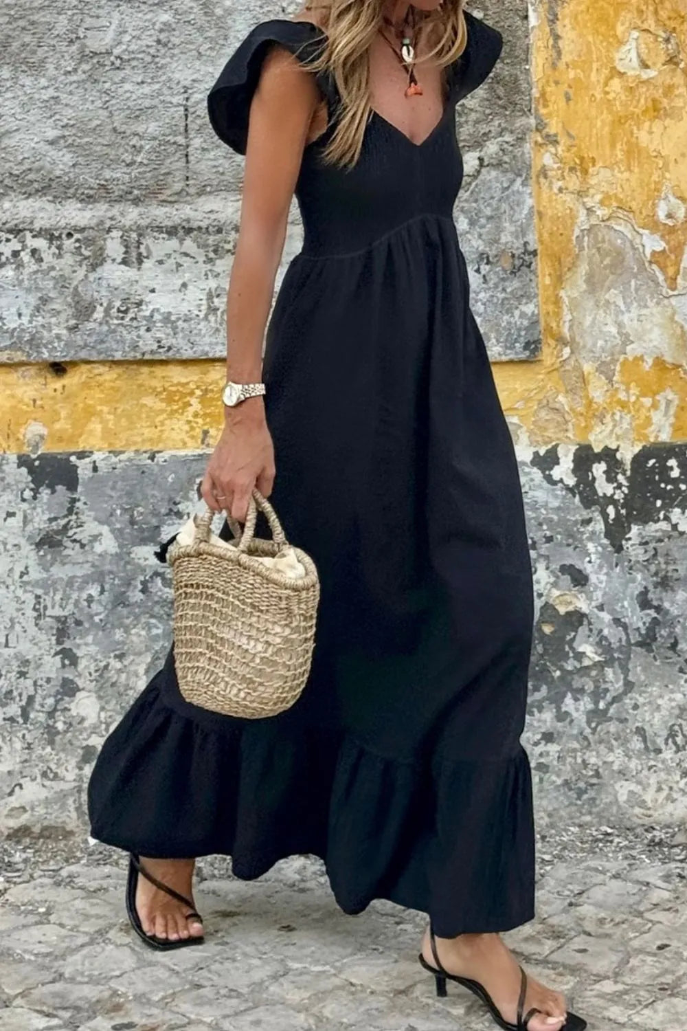 V Neck Ruffled Cap Sleeve Smocked Maxi Dress model wearing black dress and carrying a woven bag against a colorful wall.