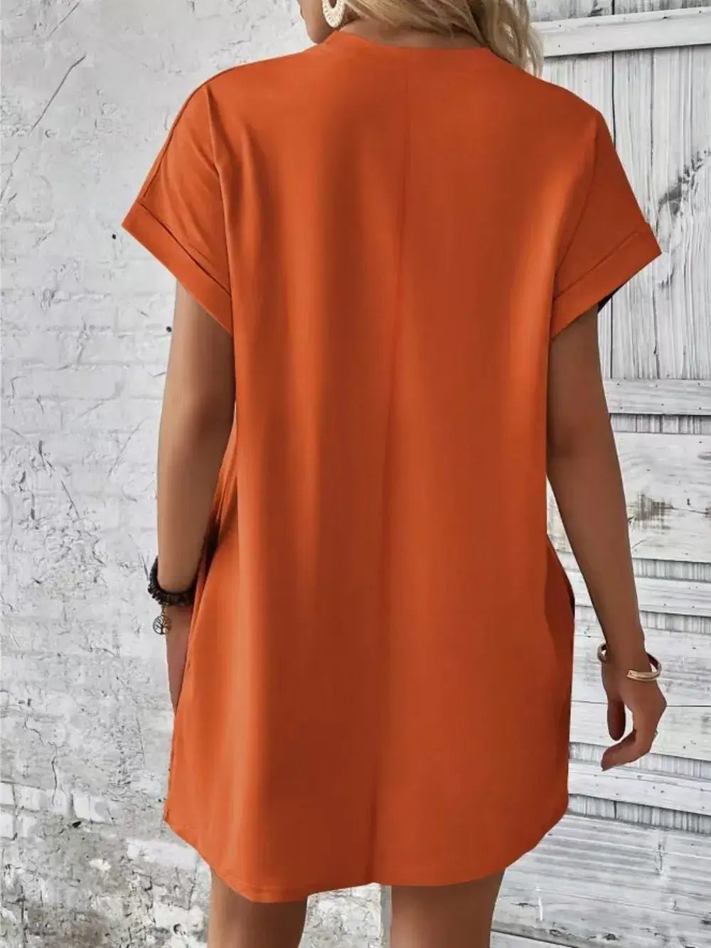 Back view of a woman wearing an orange Pocketed Round Neck Short Sleeve Dress against a rustic white wall.