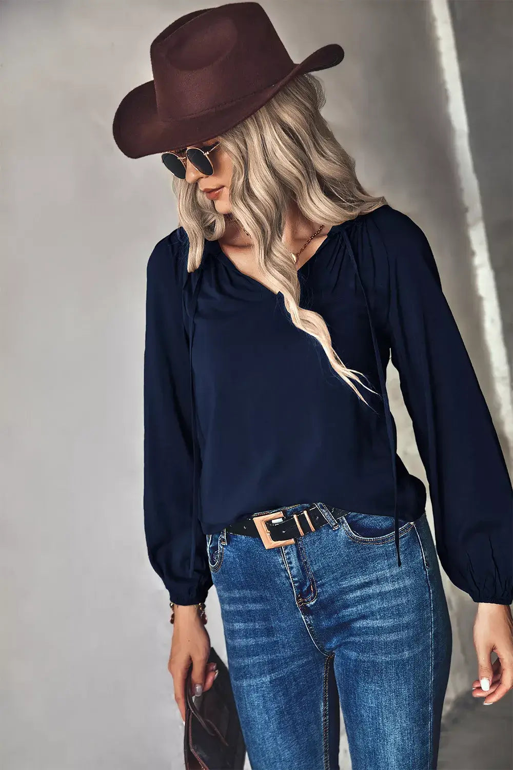 Woman wearing a tie neck long sleeve blouse with blue jeans, complemented by a brown hat and sunglasses.