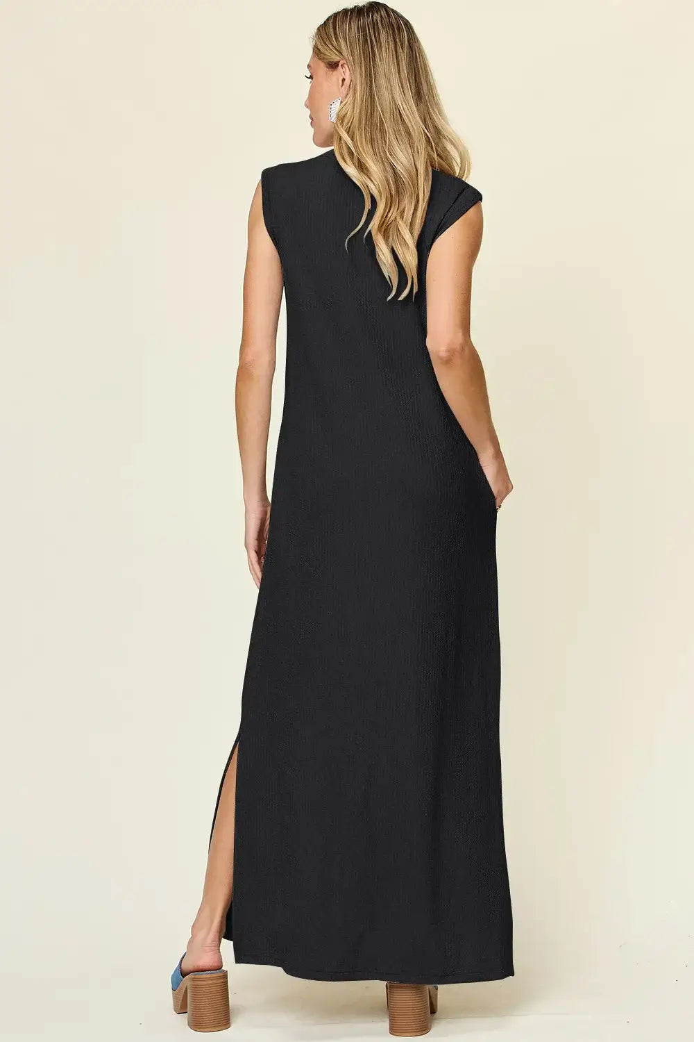 Back view of a black sleeveless maxi dress with a mock neck and side slit, showcasing its sleek and modern design.