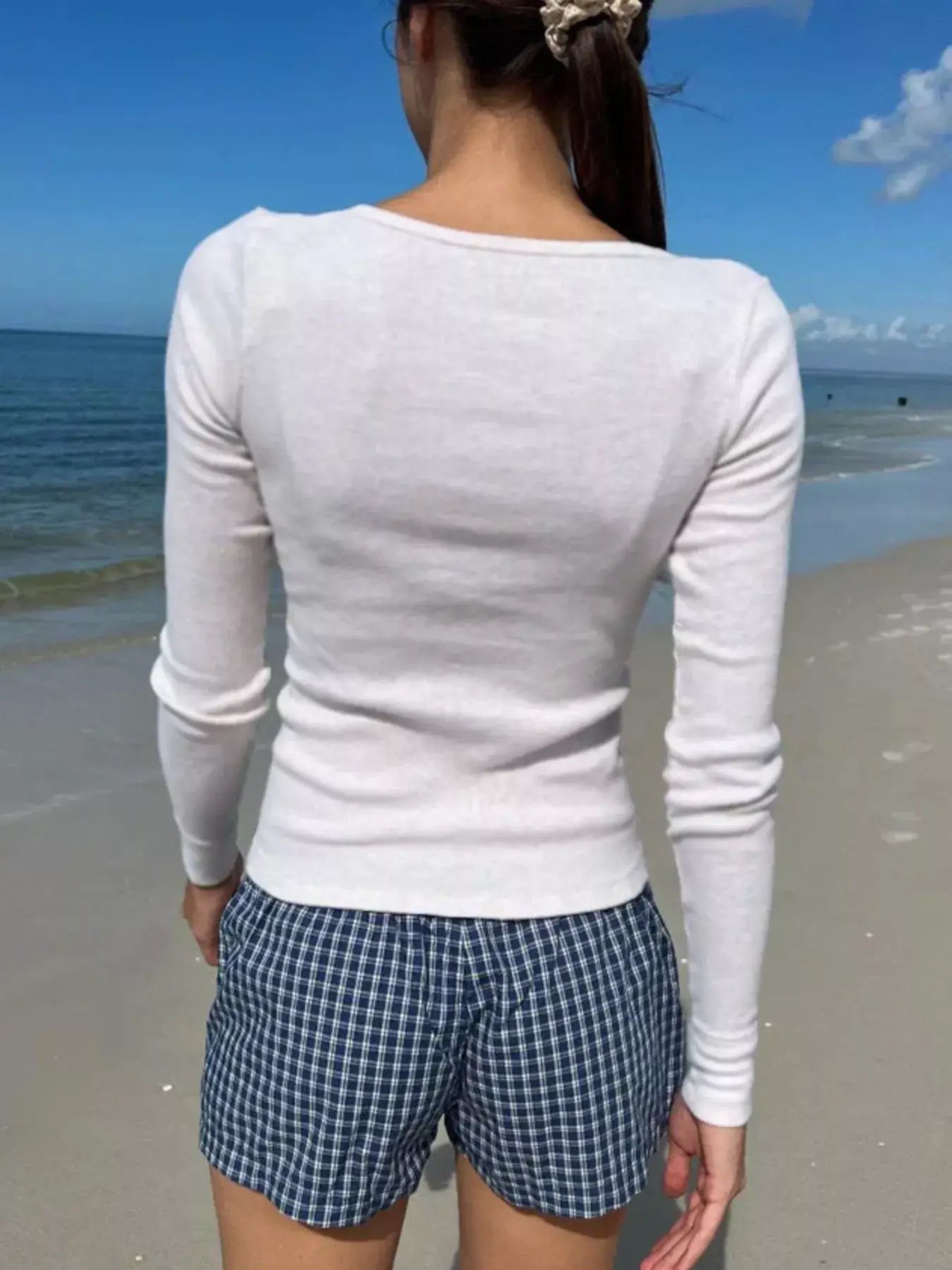 Back view of a woman wearing a white long sleeve T-shirt with checkered shorts on a beach.