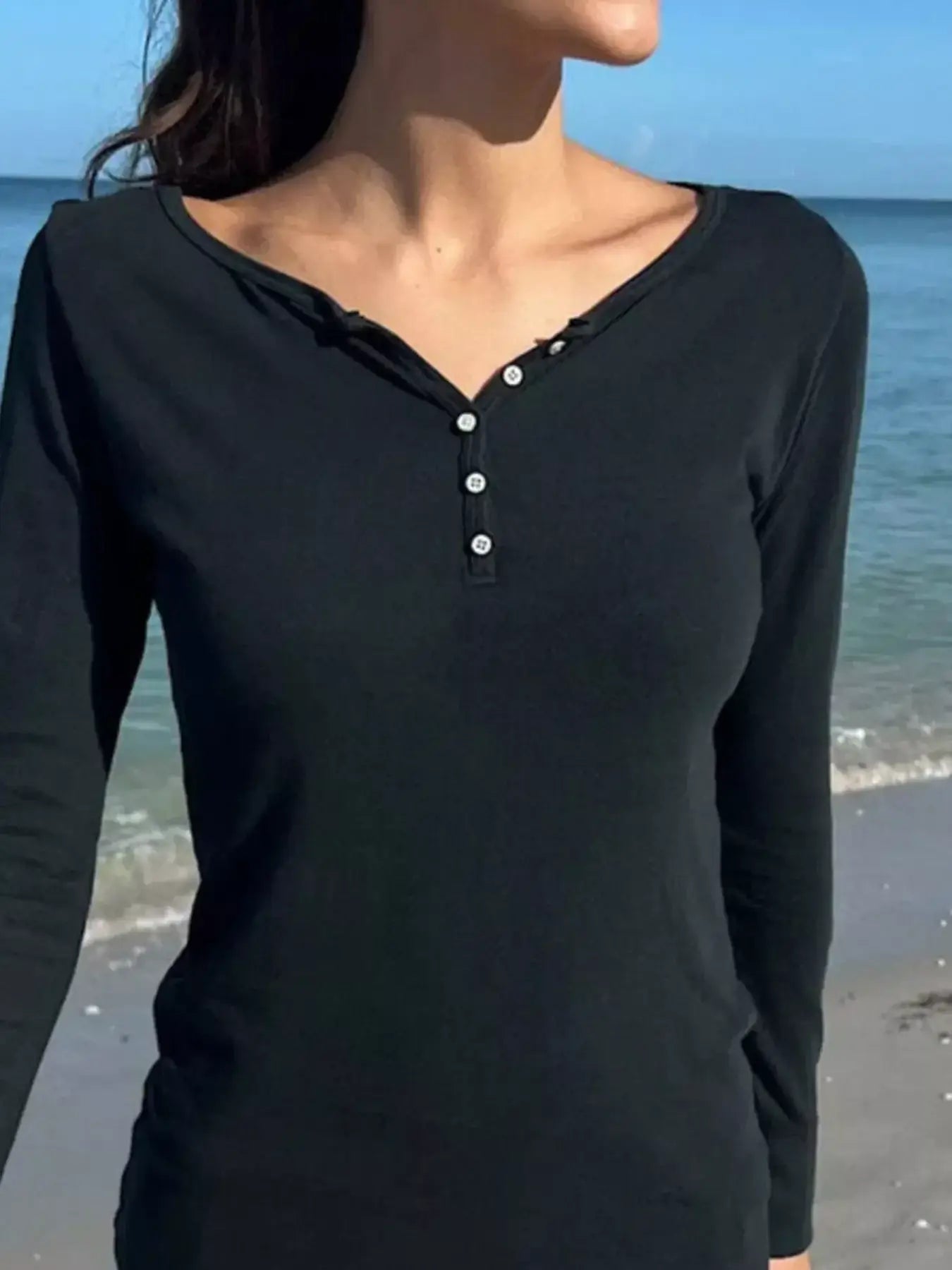 Model wearing a black long sleeve Henley T-shirt with buttoned design at the beach.