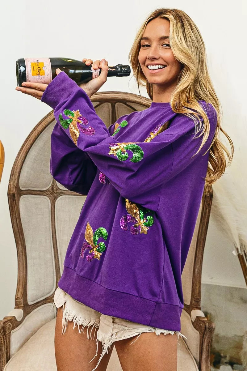 BiBi Fleur De Lis Sequin Patches Mardi Gras Sweatshirt worn by a woman holding champagne in a stylish setting.