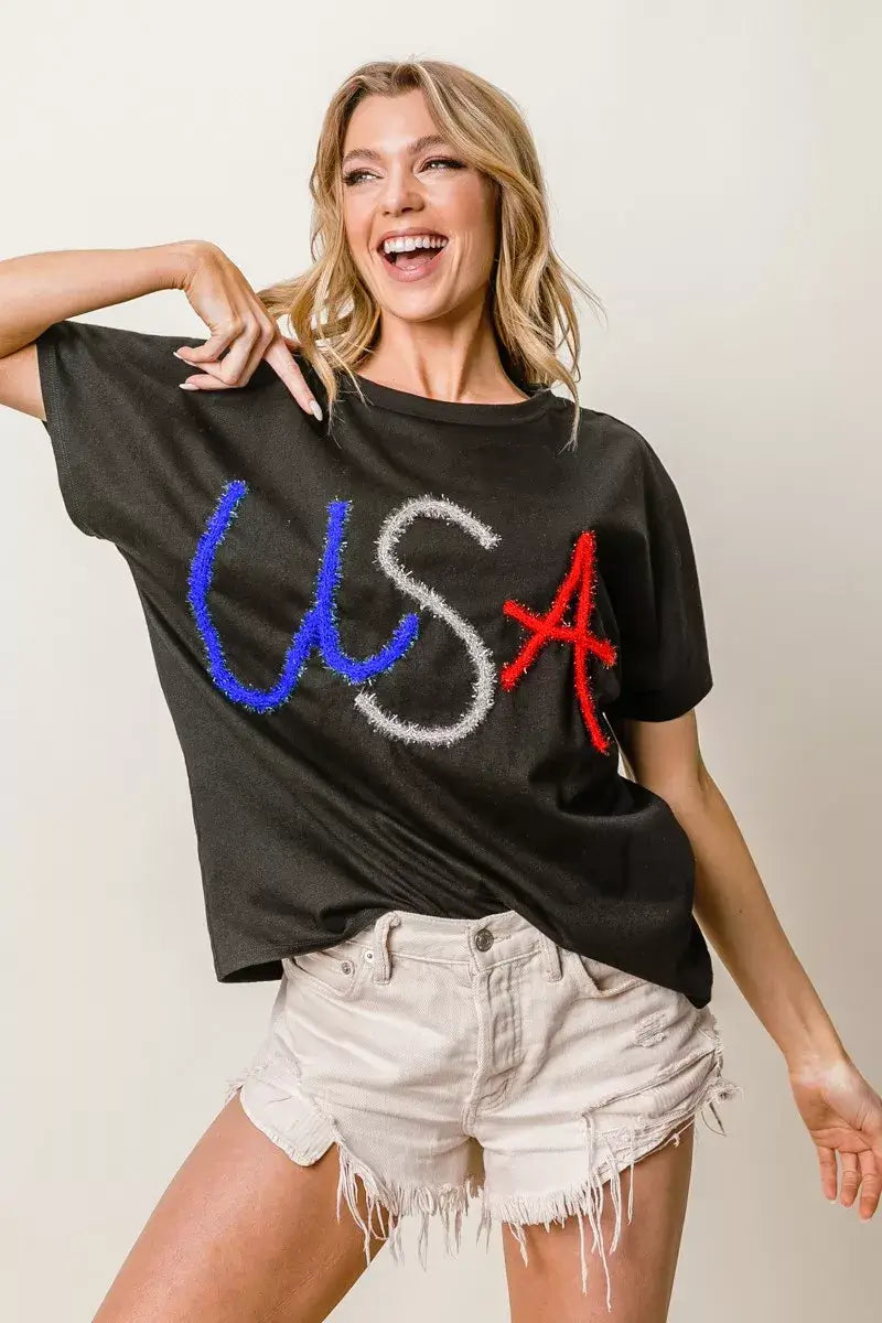 Woman wearing a black dolman sleeve top with 'USA' in rhinestones, styled with white shorts, showcasing a relaxed and stylish look.