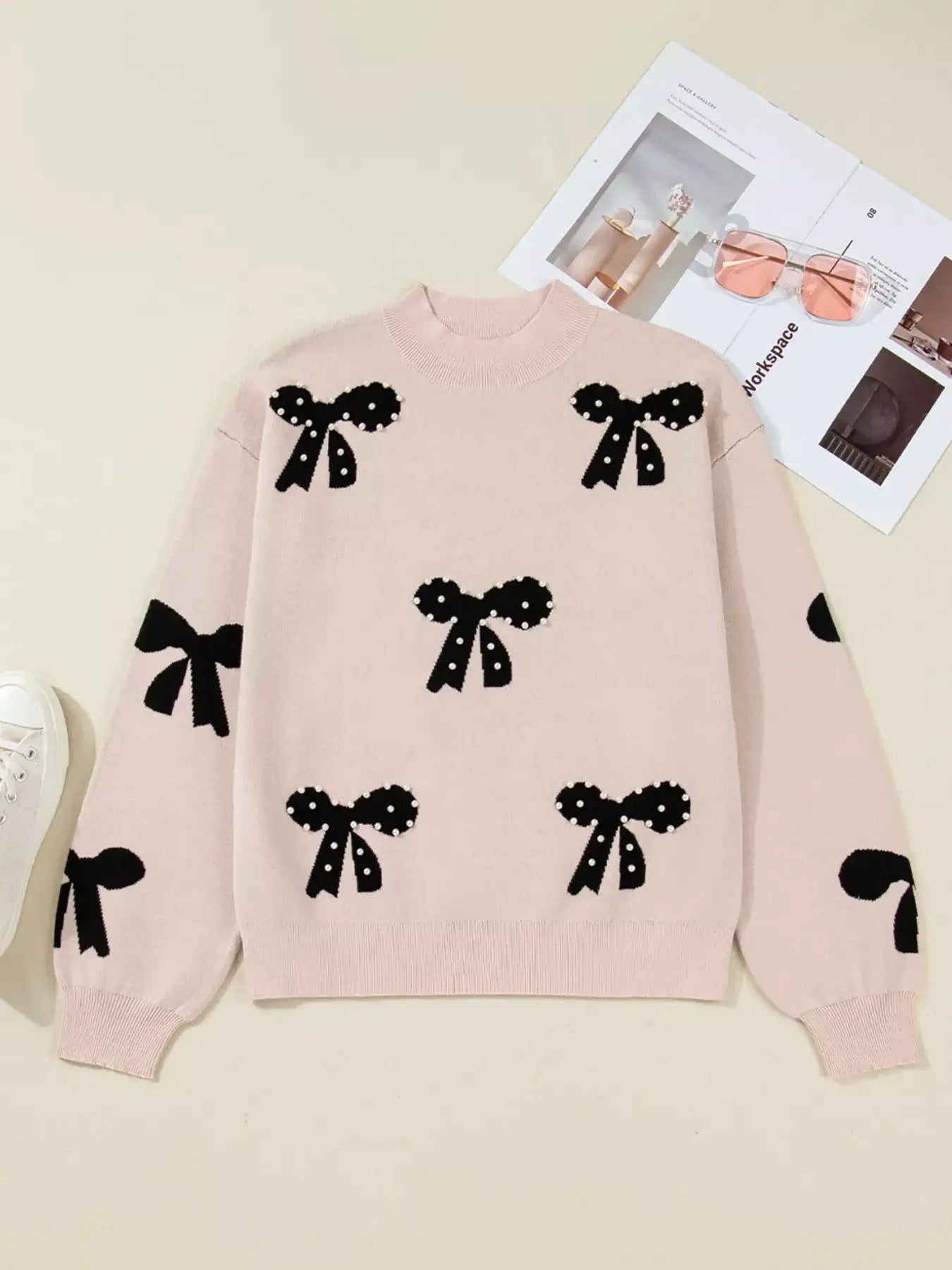 Pearl embellished bow pattern loose sweater in pink with black bow designs and pearl accents on a flat surface.