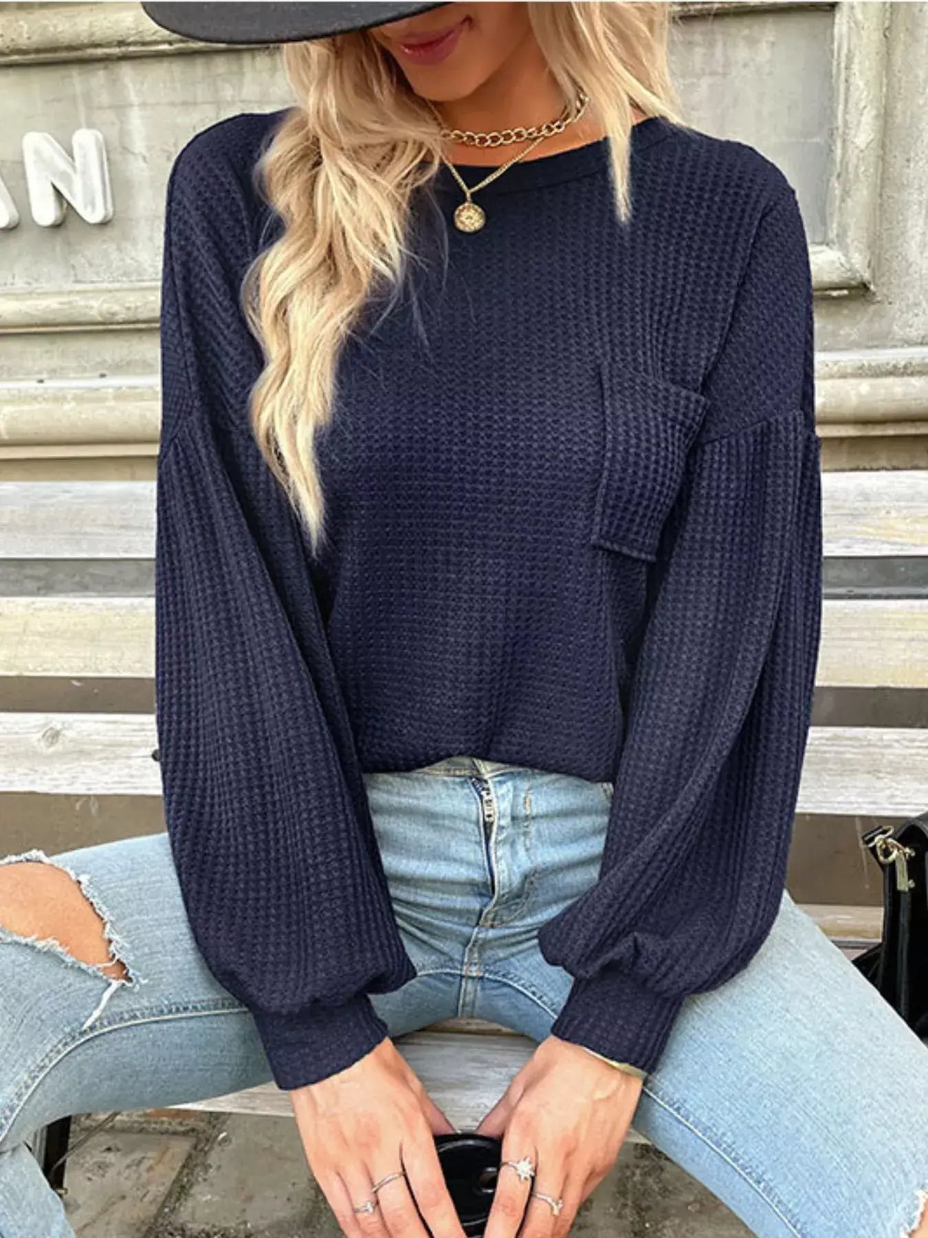 Woman wearing a navy waffle knit round neck top with long sleeves, paired with light blue jeans, sitting on a bench.