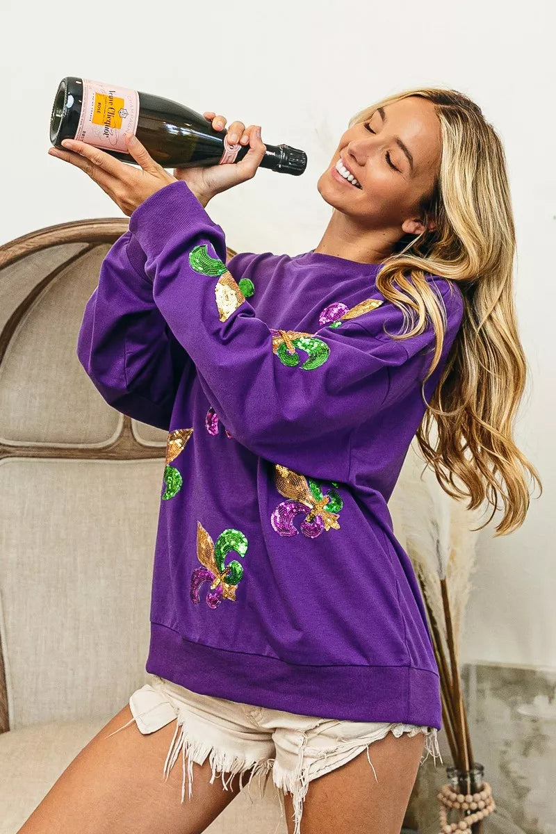 Model wearing BiBi Fleur De Lis Sequin Patches Mardi Gras Sweatshirt, celebrating with a bottle of champagne.