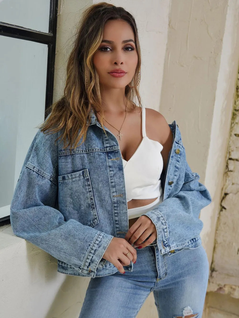 Woman wearing a Button Down Drop Shoulder Denim Jacket over a white top, posing by a window.
