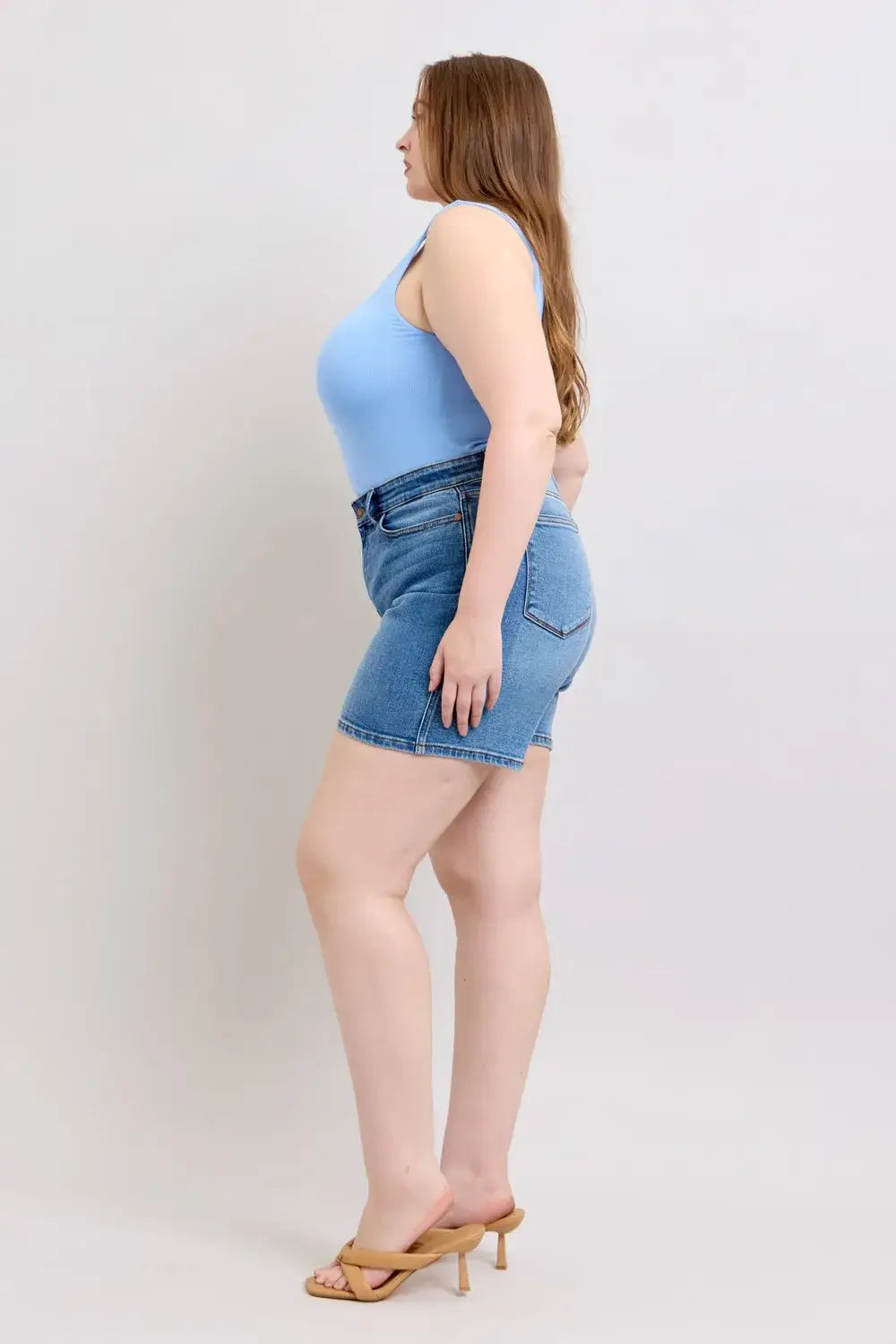Model wearing Judy Blue Plus Size Denim Shorts in blue with a fitted tank top and sandals, showcasing a side profile.