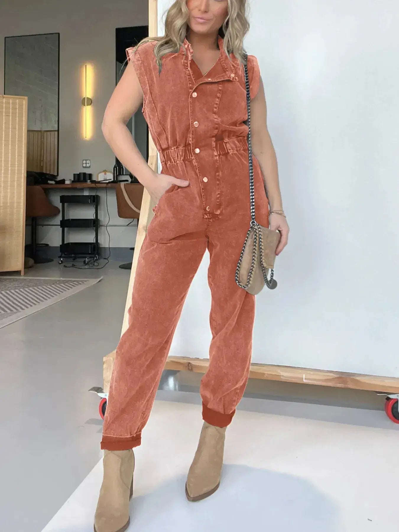 Model wearing a rust-colored denim jumpsuit with cap sleeves and a belted waist, paired with ankle boots.