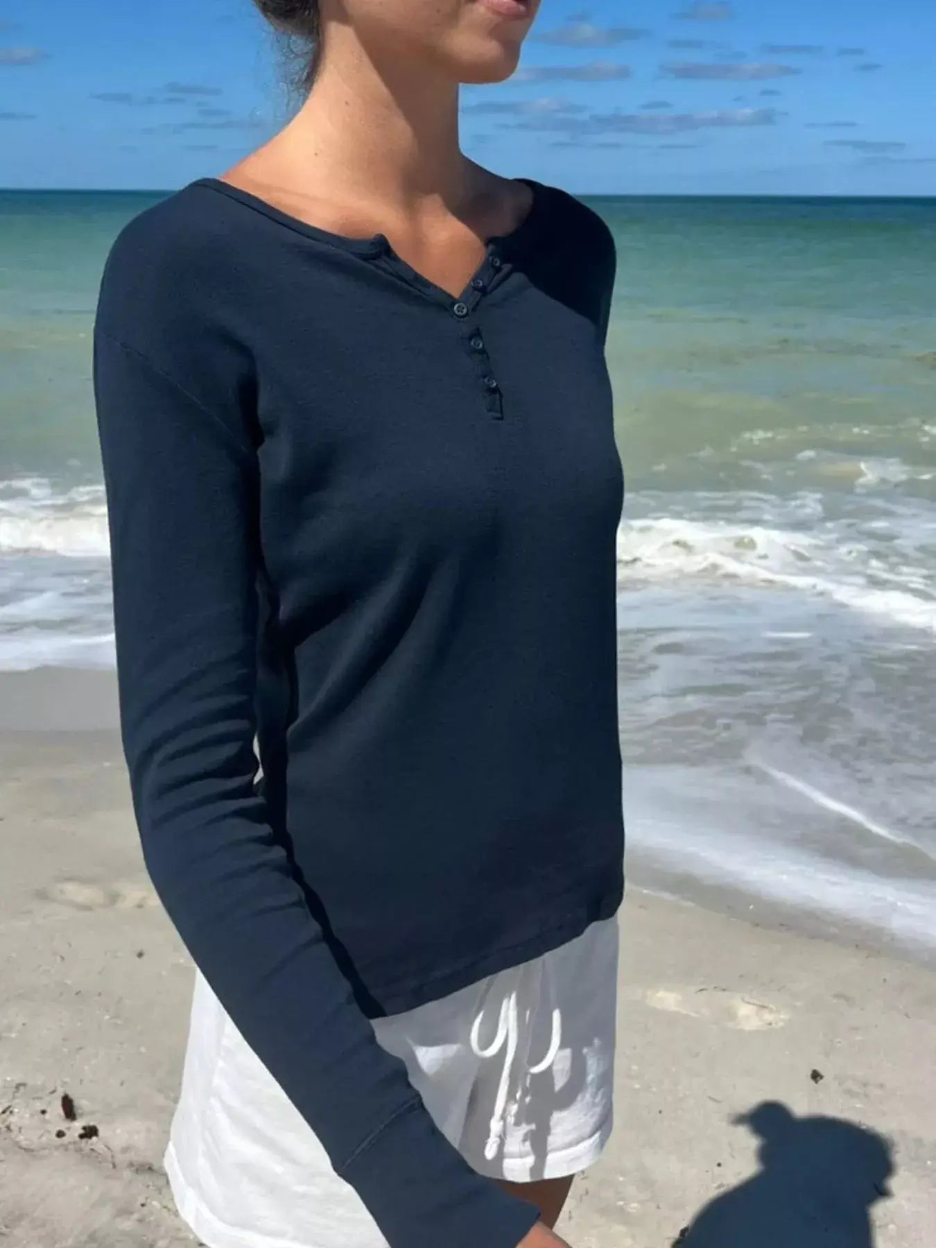 Model wearing a navy striped long sleeve Henley T-shirt at the beach with ocean in the background.