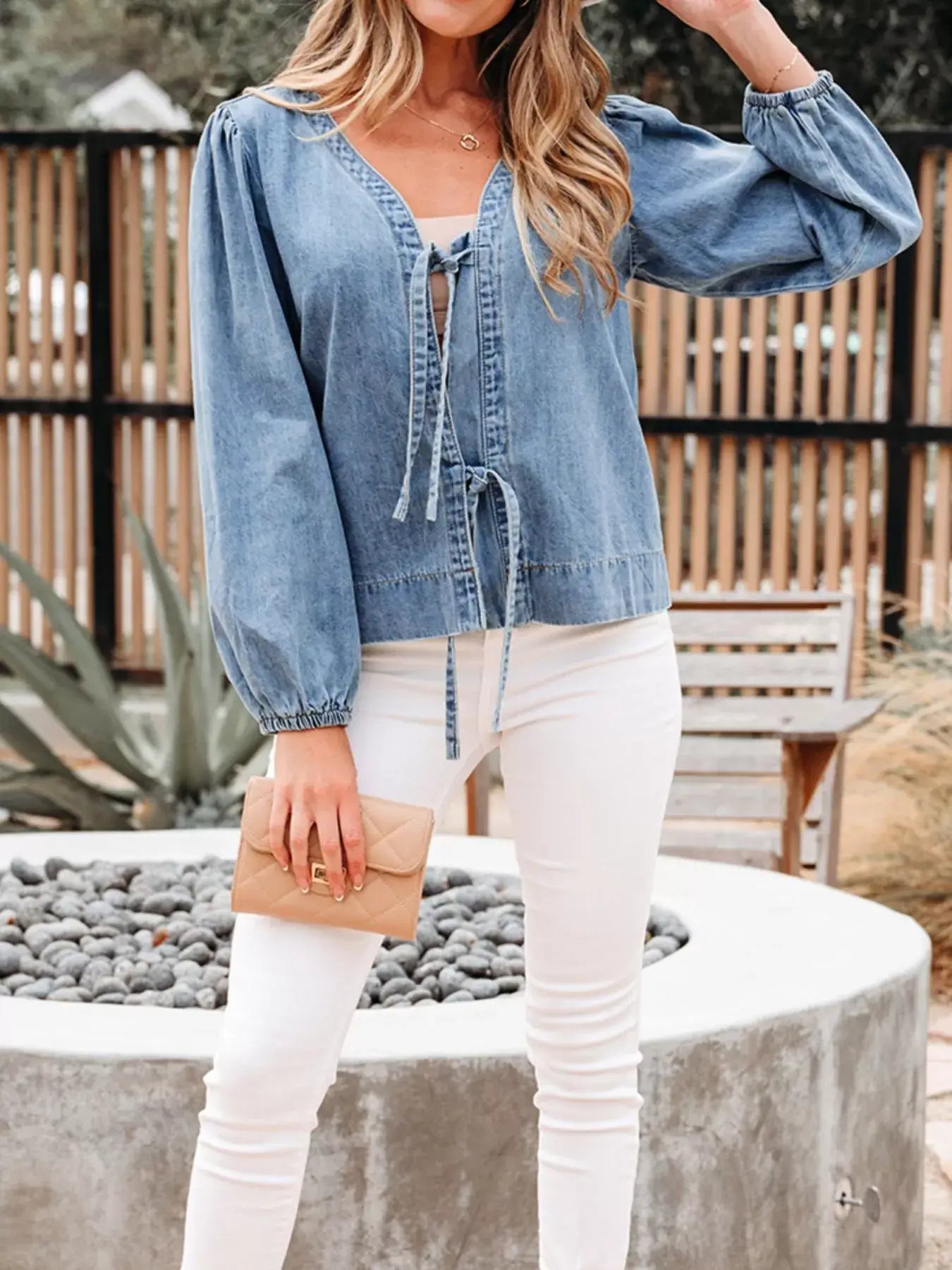 Woman wearing a tied front puff sleeve denim top with dramatic sleeves, paired with white jeans and holding a beige clutch.
