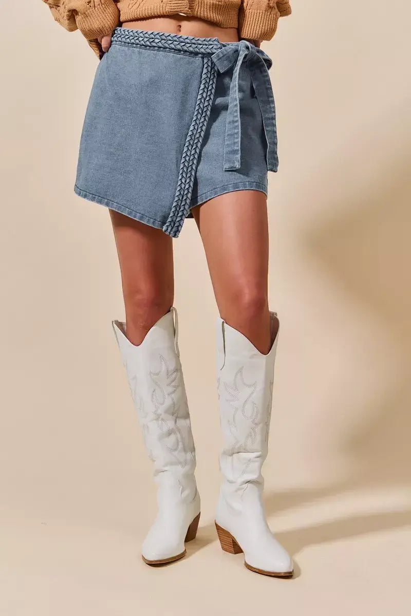 Model wearing a wrap denim skort with braided trim and white cowboy boots, showcasing a trendy casual style.