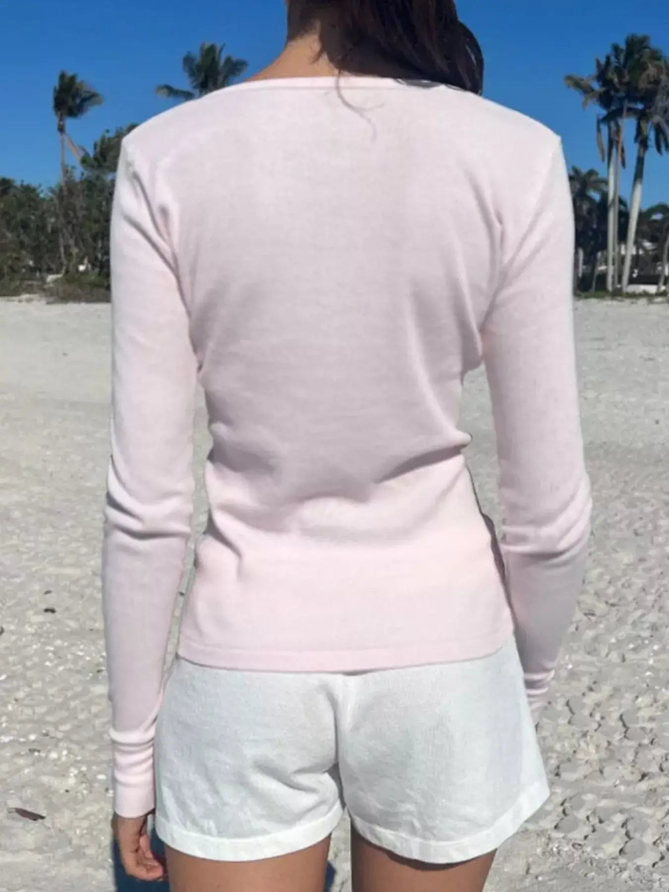 Woman wearing a pale pink striped long sleeve Henley t-shirt and white shorts on a beach.