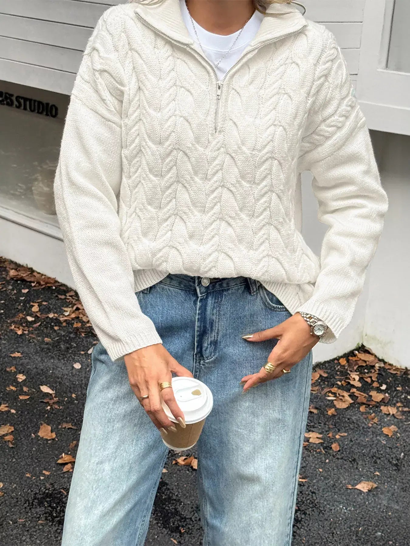 Woman in zip collar cable-knit sweater holding a coffee cup outdoors, showcasing stylish and cozy winter fashion.