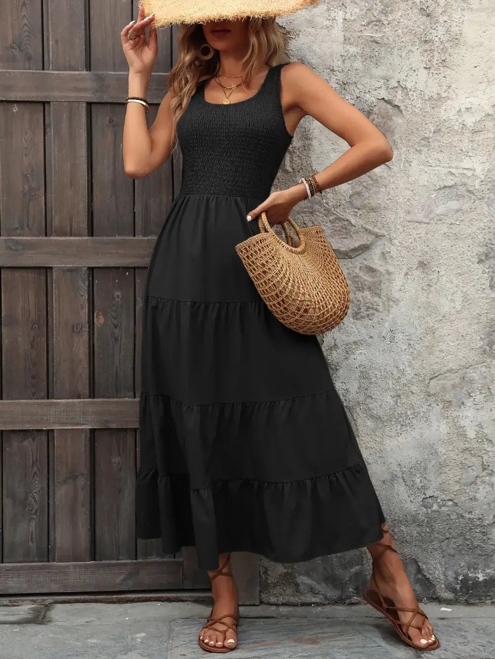 Model wearing an Ivy Lane Smocked Scoop Neck Sleeveless Tank Dress in black, holding a straw bag against a wooden backdrop.