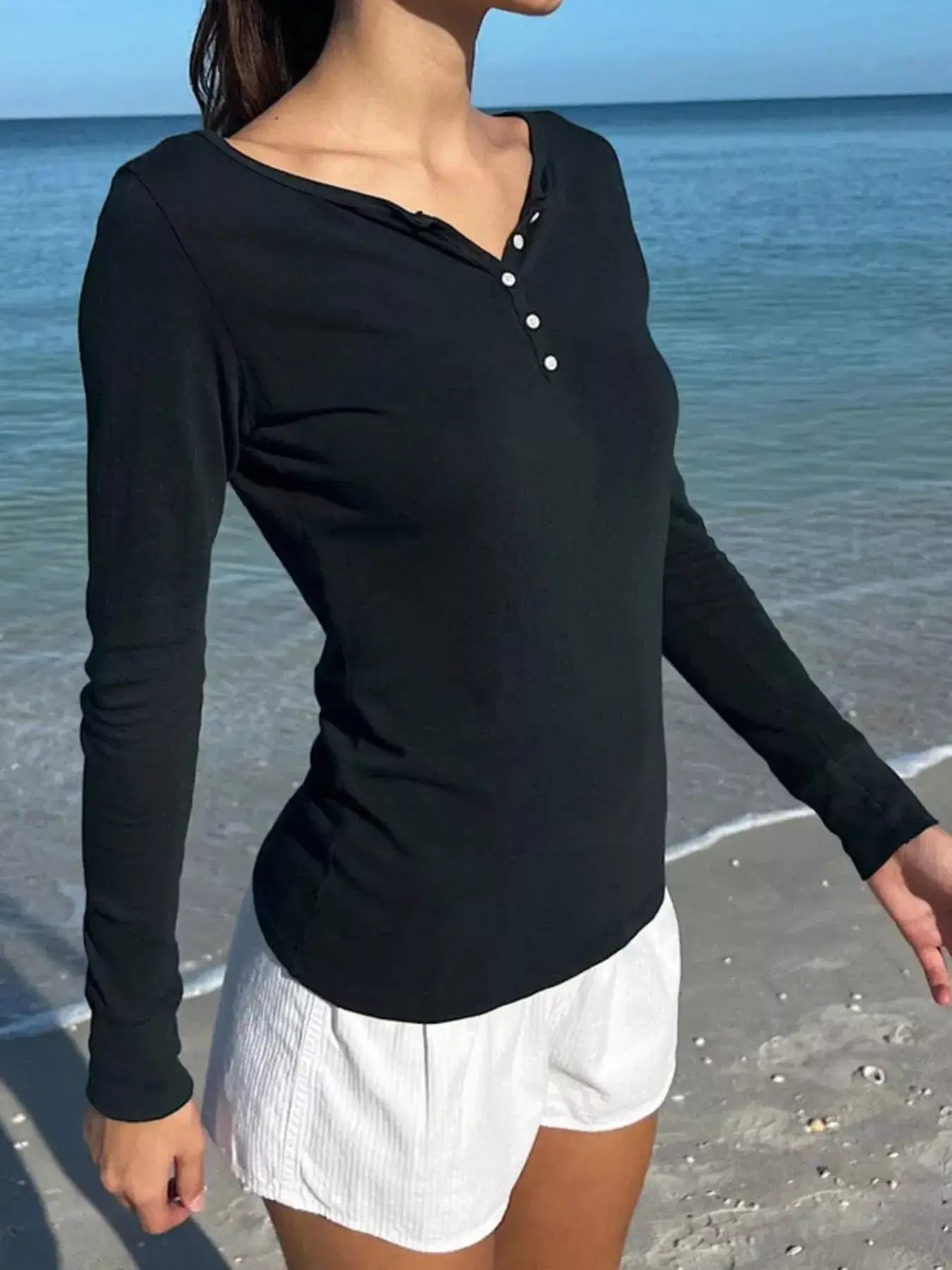 Woman wearing a black long sleeve henley t-shirt by the beach, styled with white shorts.