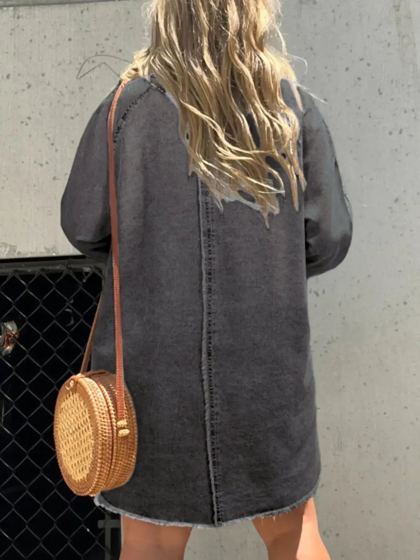 Back view of a woman wearing a long sleeve denim shirt dress, paired with a round woven bag, against a textured background.