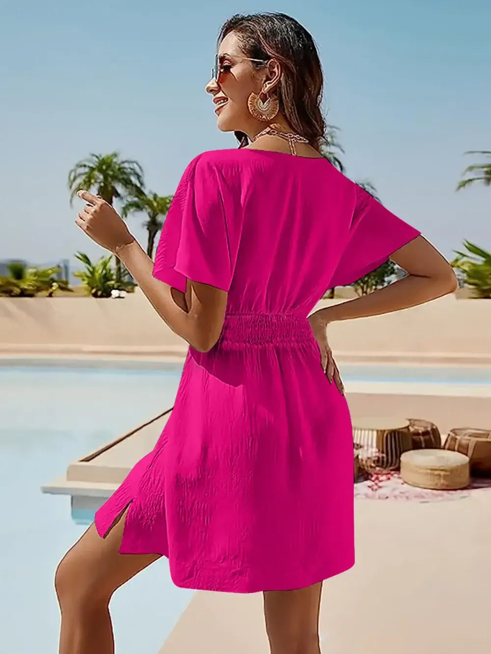 Woman wearing a bright pink smocked V-neck short sleeve dress by a poolside, showcasing a stylish slit and elegant silhouette.