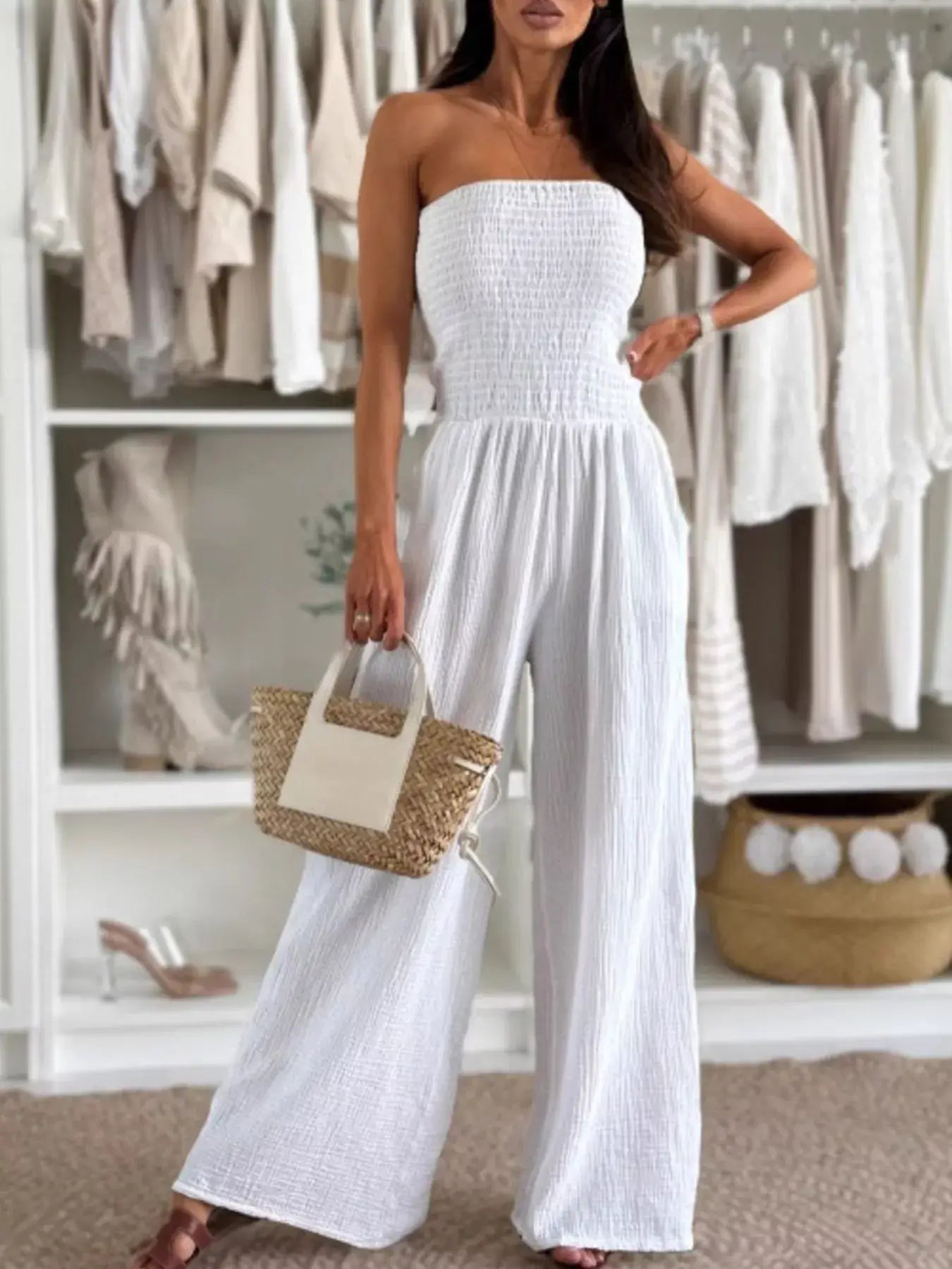 Model wearing a Smocked Tube Wide-Leg Jumpsuit in white, holding a straw bag, styled against a backdrop of neutral clothing.