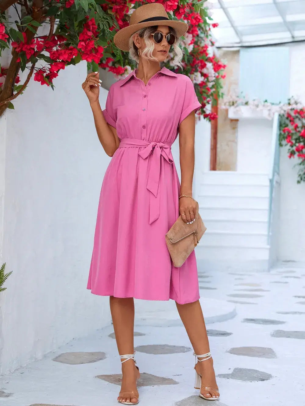 Model wearing a pink buttoned tie waist short sleeve dress with sunglasses and a hat, standing in a floral setting.