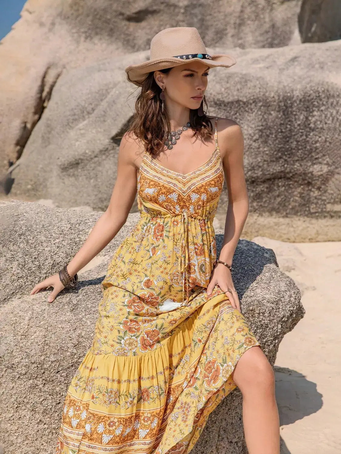 Model wearing a Tassel Printed Tiered Cami Dress with bohemian vibe, sitting on rocks at the beach