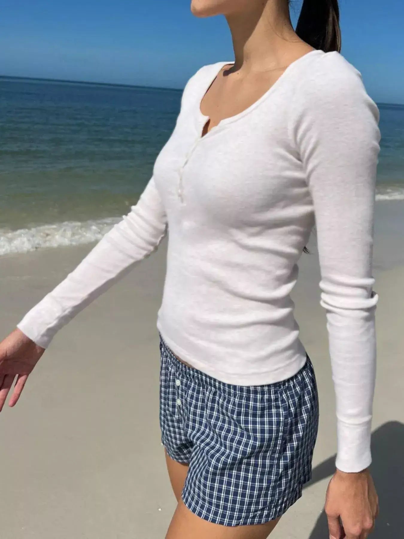 Model wearing a white striped long sleeve Henley T-shirt by the beach, paired with shorts on a sunny day.