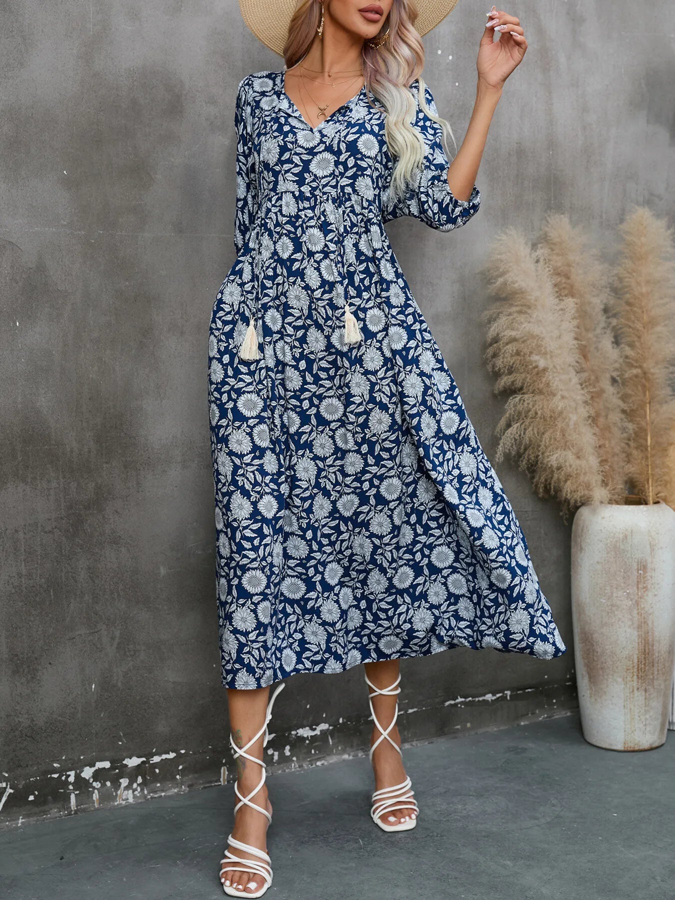 Model wearing a Printed Tie Neck Midi Dress featuring floral print and tassels, styled with a wide-brimmed hat and sandals.