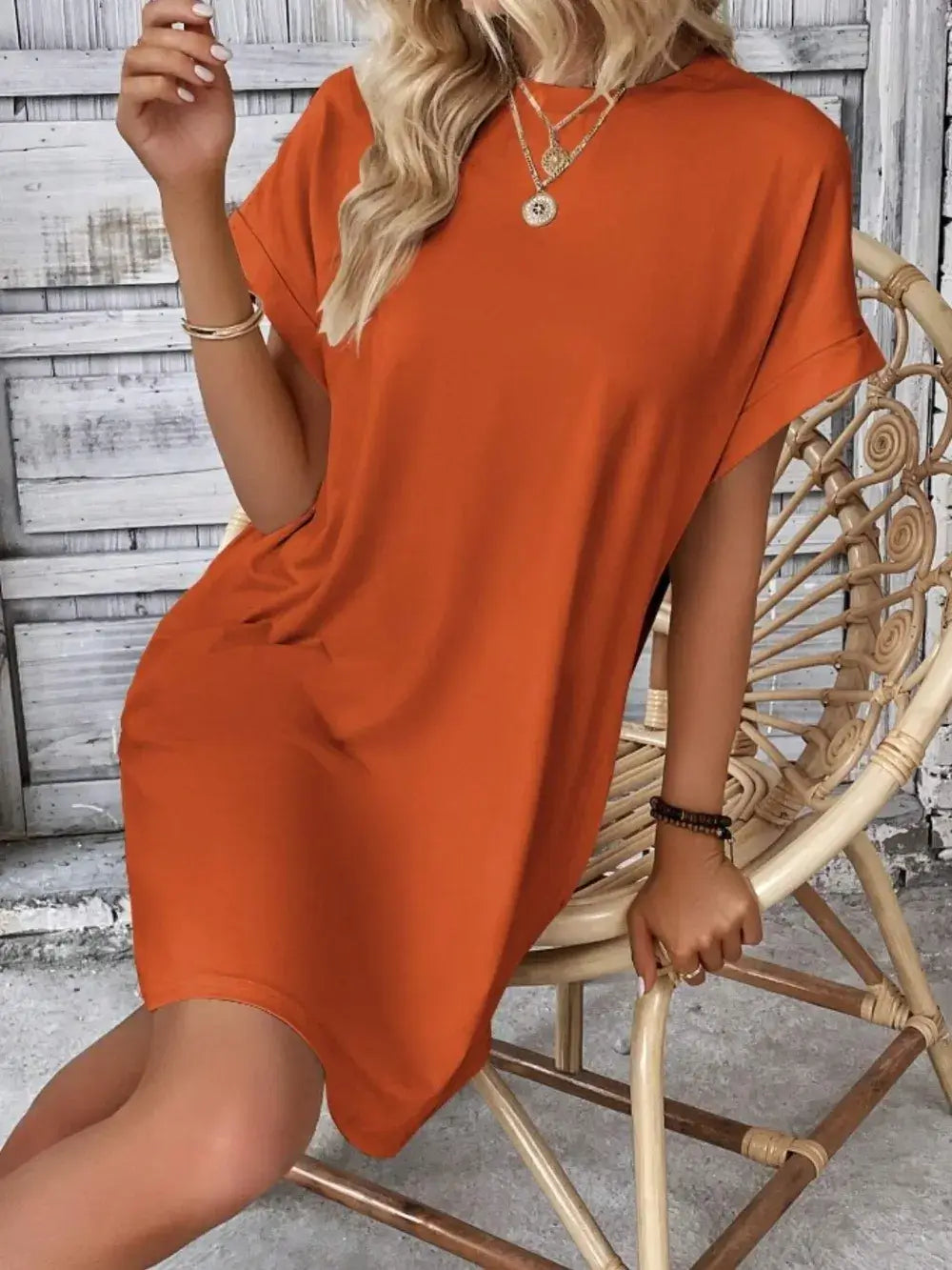 Casual pocketed round neck short sleeve dress in vibrant orange, worn by a model seated on a rattan chair.