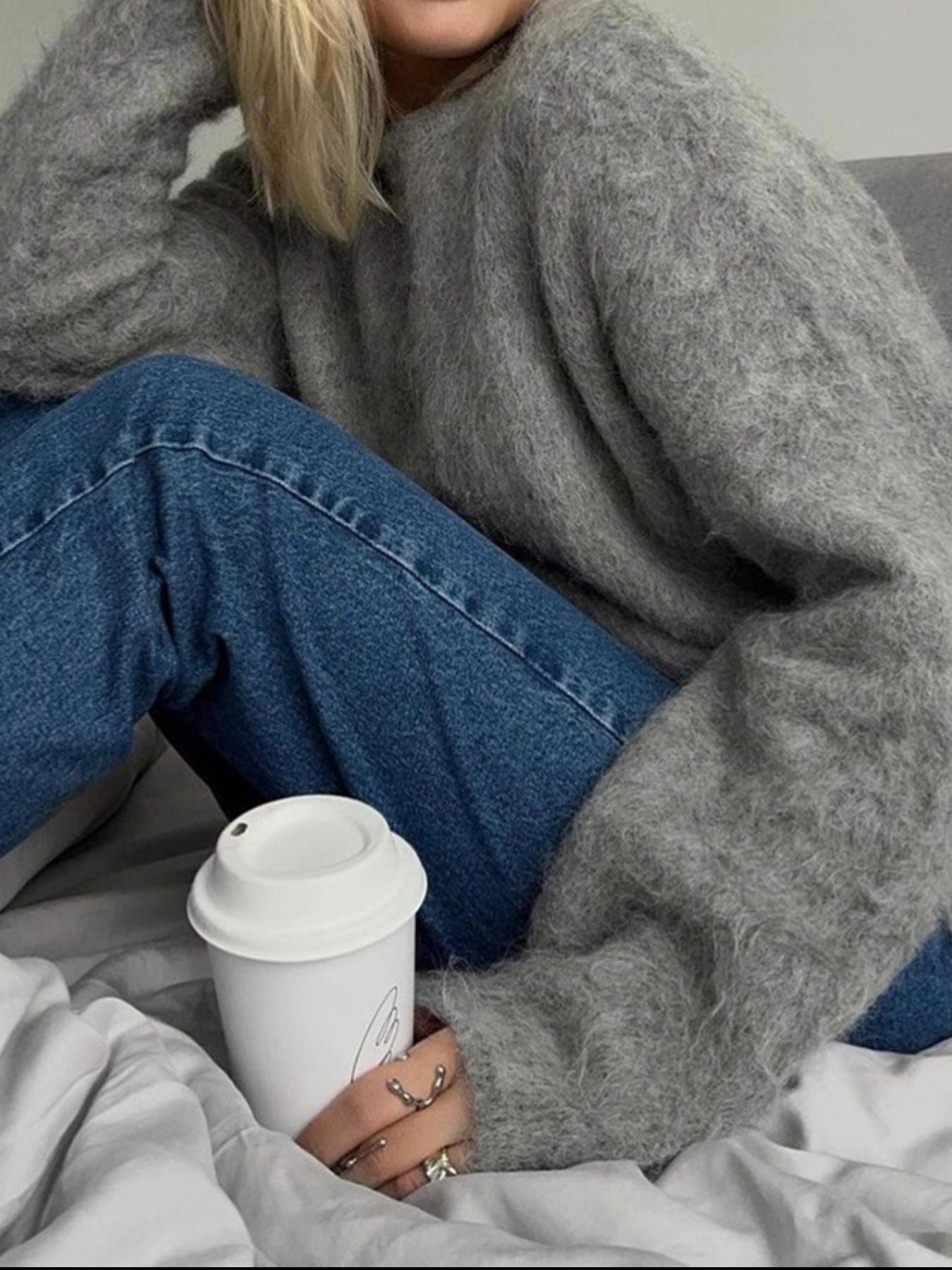 Woman wearing a Fuzzy Round Neck Long Sleeve Sweater, holding a coffee cup, seated on a bed with blue jeans.