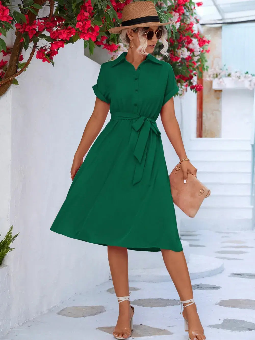 Woman in a green buttoned tie waist short sleeve dress, standing outdoors with floral backdrop.