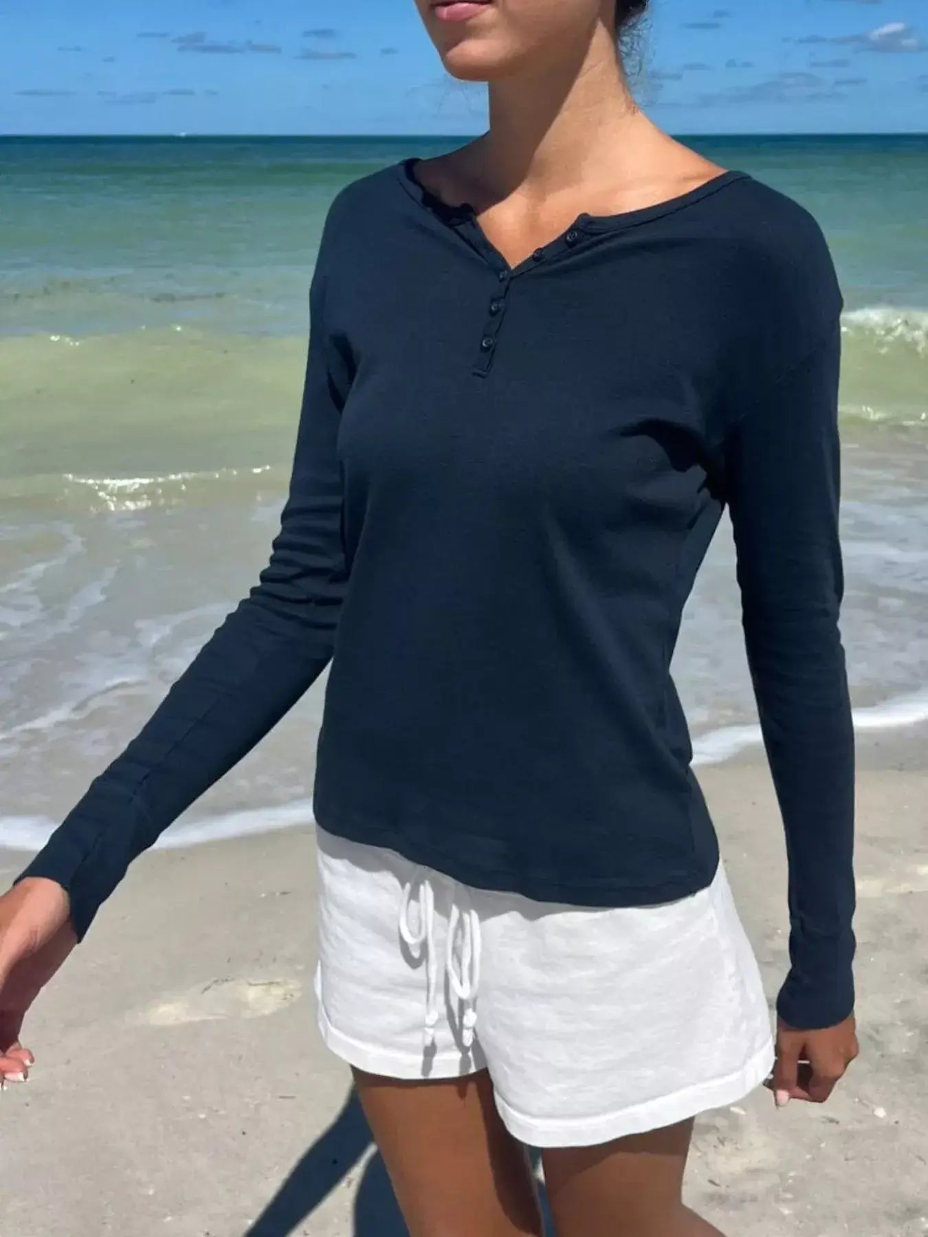 Model wearing a navy striped long sleeve Henley t-shirt at the beach, with white shorts and ocean background.