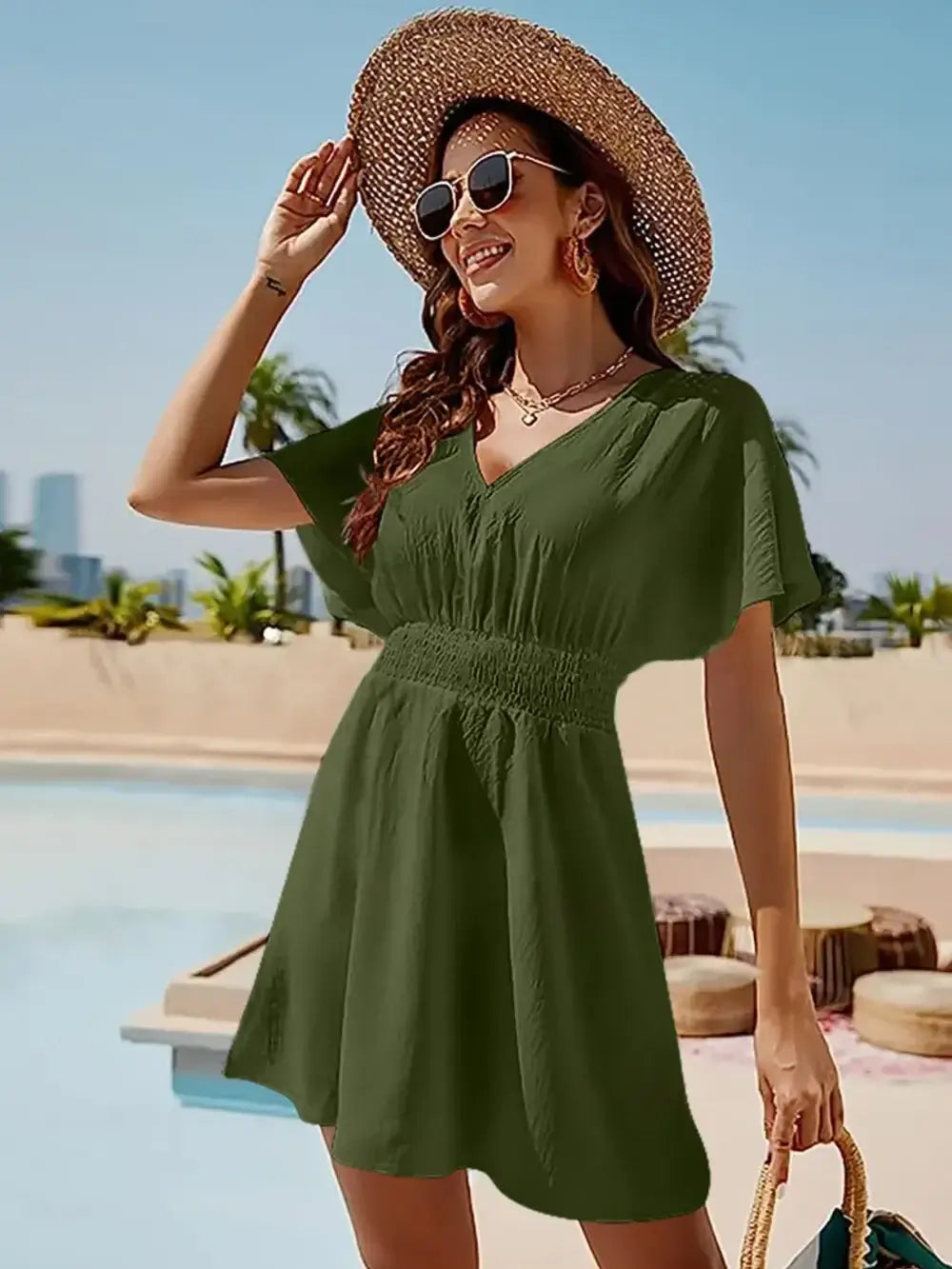 Woman wearing a smocked V-neck short sleeve dress in olive green, stylishly accessorized by a sun hat and sunglasses by the pool.