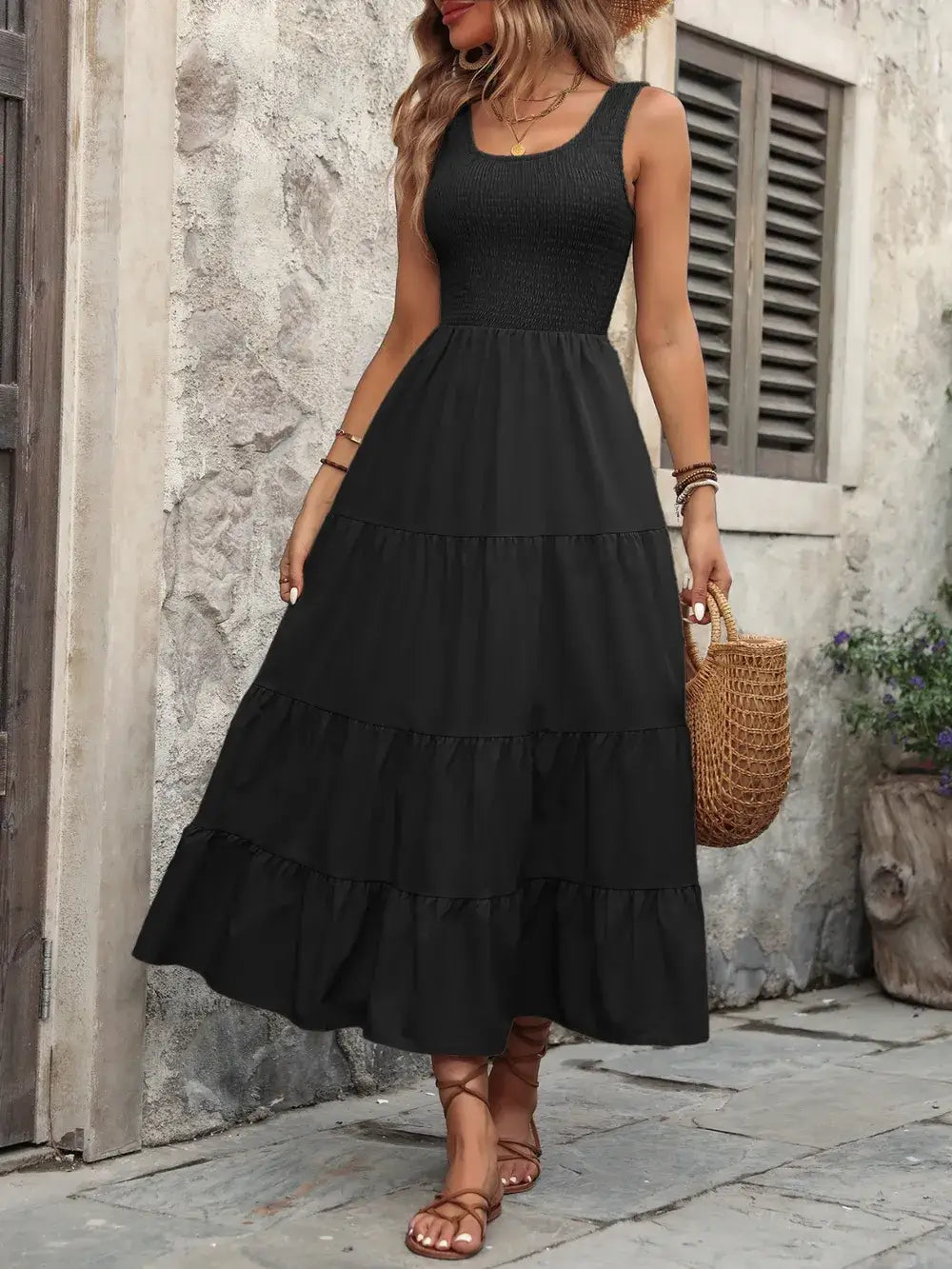 Woman in a black smocked scoop neck sleeveless tank dress with a tiered skirt and a woven bag, standing outdoors.