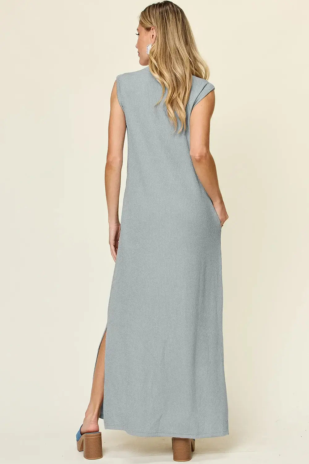 Back view of a woman wearing a sleeveless gray maxi dress with a mock neck and side slit.